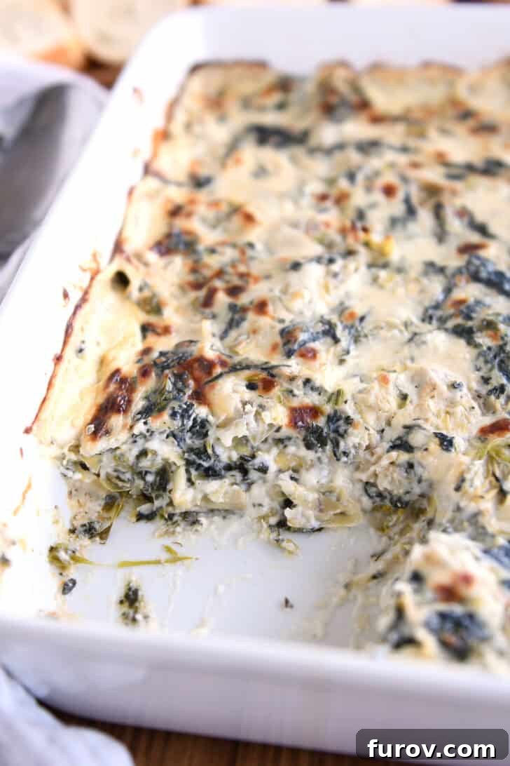 Ultimate Creamy Spinach Artichoke Dip 7 A white casserole dish with a creamy cooked spinach artichoke dip, perfectly golden. A scoop is taken out of the corner of the dish, revealing its rich texture.