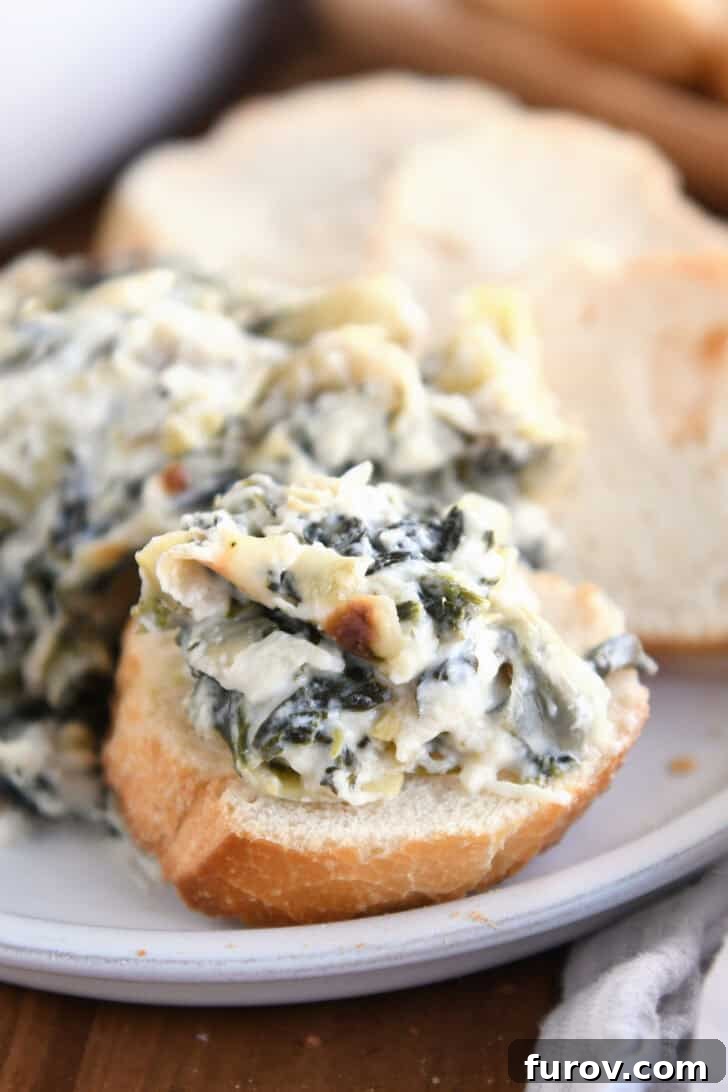 Ultimate Creamy Spinach Artichoke Dip 8 A small piece of sliced baguette with creamy spinach artichoke dip on top, garnished slightly. More sliced bread and the full dip dish are in the background, inviting a delightful experience.