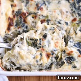 Ultimate Creamy Spinach Artichoke Dip 9 A large spoonful of creamy spinach artichoke dip being taken out of a white casserole dish full of the dip, highlighting its texture and golden crust.