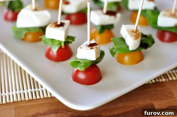 White platter with halved cherry tomatoes, a basil leaf, and a cube of fresh mozzarella skewered on toothpicks. Drizzled with balsamic glaze.