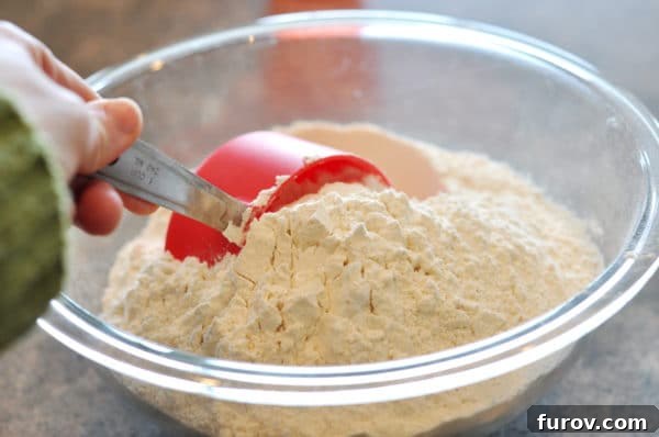 A red measuring cup scooping flour out of a glass bowl, illustrating the start of the dip and shake method.