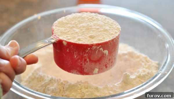 A red measuring cup full of white flour being leveled by shaking it over a glass bowl of flour.