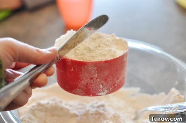 A red measuring cup full of white flour being leveled off with a butter knife, demonstrating the sweep method.
