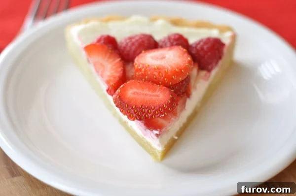 A triangle slice of fruit pizza on a white plate, showcasing the layers of tender cookie crust, creamy frosting, and fresh fruit.