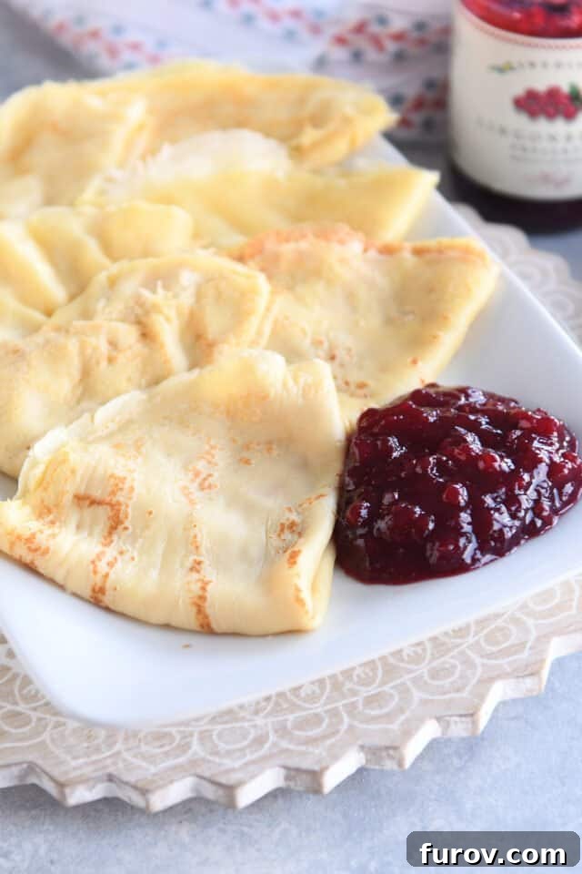 Seven perfectly folded Swedish pancakes neatly arranged on a white serving tray, accompanied by a small bowl of vibrant red lingonberry jam.