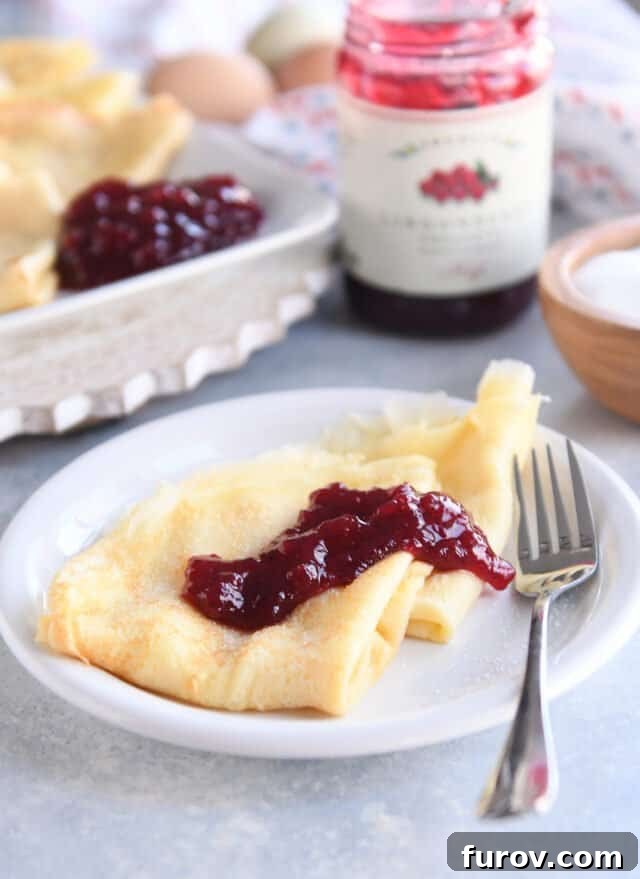 Two perfectly triangle-folded Swedish pancakes on a white plate, served with a dollop of red jam and a fork, ready to be enjoyed.