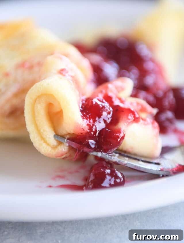 A curled up piece of Swedish pancake delicately placed on a fork, topped with a rich red dollop of lingonberry jam, ready for a bite.