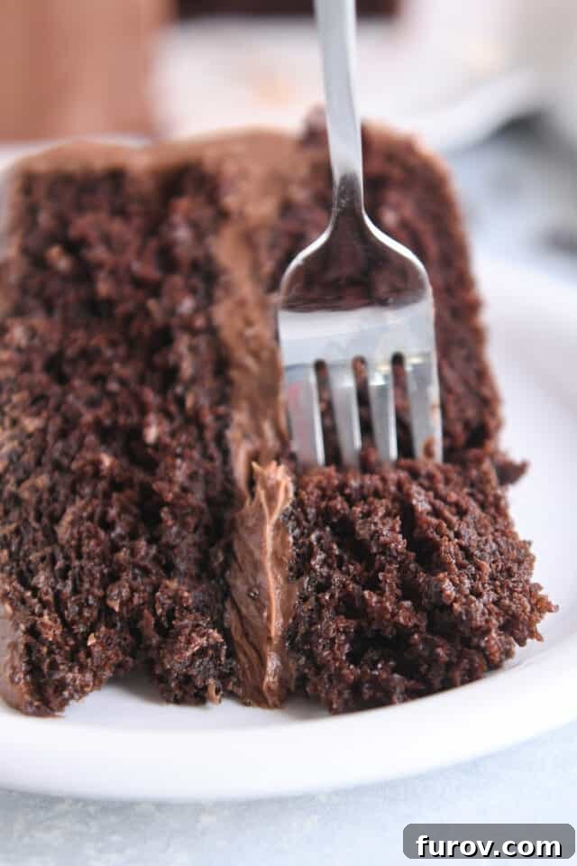 A fork gently scoops a perfect bite of chocolate cake, showcasing its tender, moist interior and luscious frosting, on a white plate.
