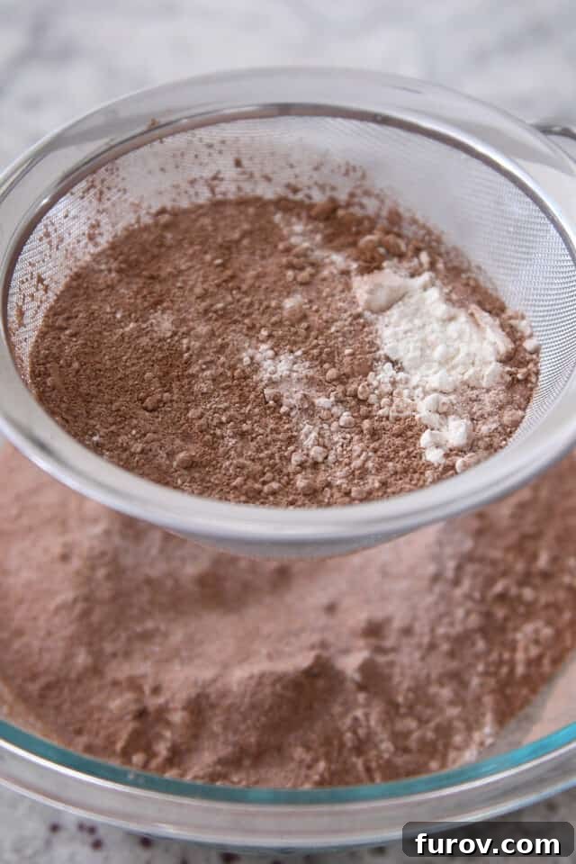 A fine mesh strainer sifts cocoa powder, flour, and sugar into a clear glass bowl, illustrating the essential first step for a smooth chocolate cake batter.