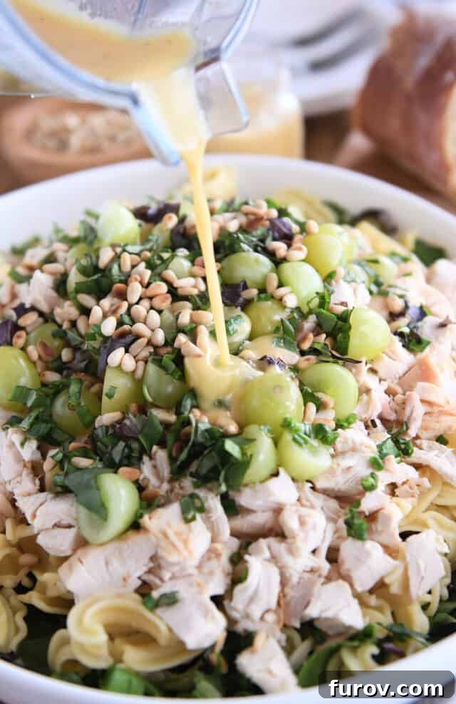 Close-up shot of vibrant vinaigrette dressing being poured over a bowl of Vineyard Chicken and Pasta Salad, highlighting the fresh greens, halved grapes, and cooked chicken.