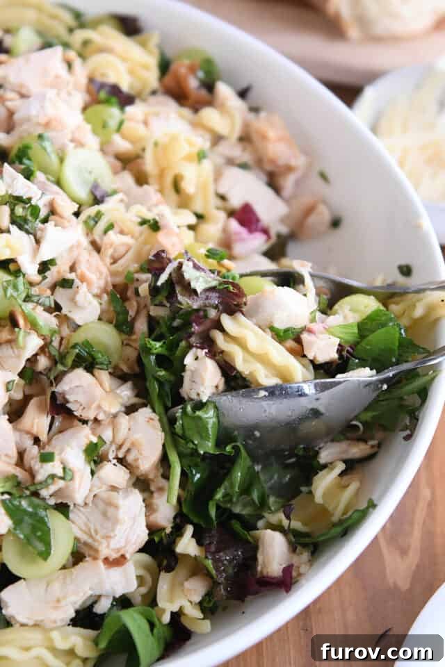 Salad tongs scooping a generous portion of Vineyard Chicken and Pasta Salad from a large white serving bowl, showcasing the mix of ingredients.