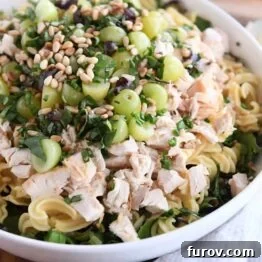 Large white bowl filled with greens, cooked pasta, chicken pieces, red and green grapes, fresh basil leaves, and toasted pine nuts, all ready for the dressing.