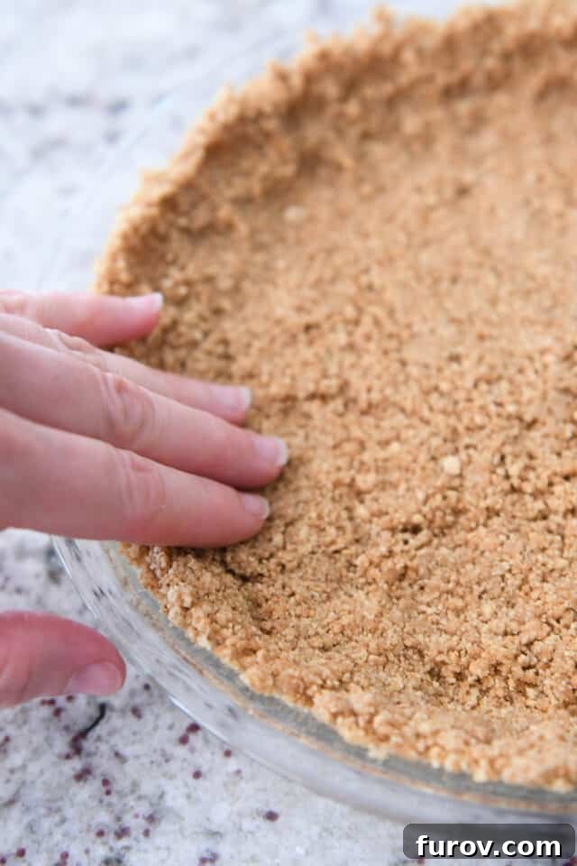 Crisp, Buttery Graham Cracker Crust 6 Fingers pressing the graham cracker crust mixture into the sides and bottom of a glass pie plate, demonstrating the initial shaping.