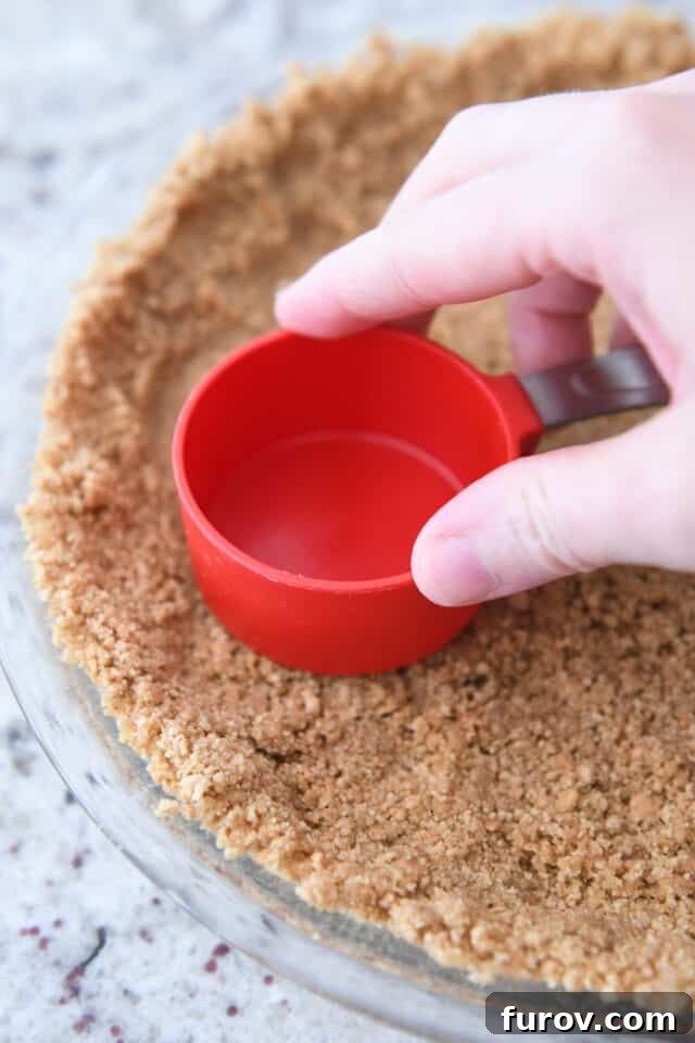 Crisp, Buttery Graham Cracker Crust 7 A measuring cup firmly pressing the graham cracker crust into a glass pie plate, ensuring a compact and even base.