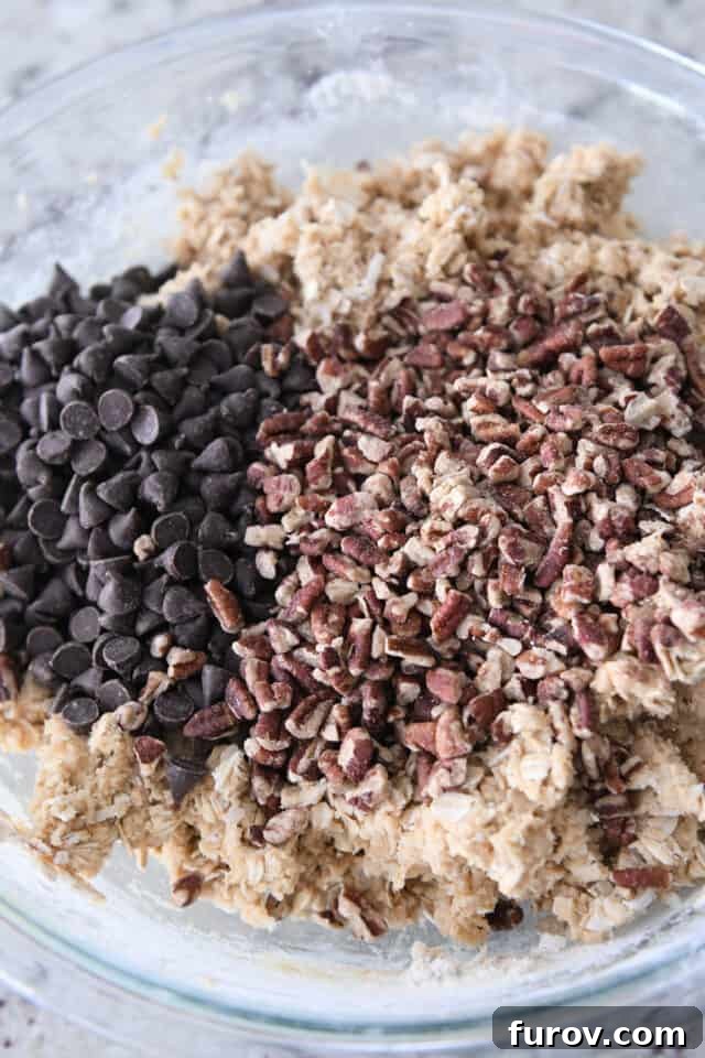 A glass bowl showcasing perfectly mixed cowboy cookie dough, generously topped with an inviting scatter of chocolate chips and toasted pecans, awaiting final incorporation.