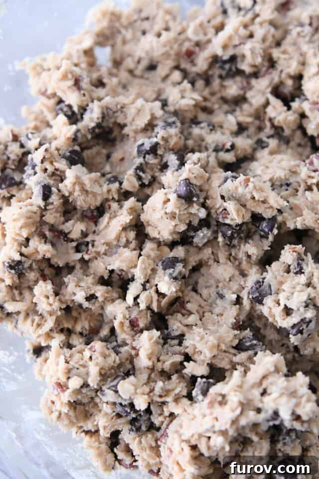 A glass bowl filled with rich cowboy cookie dough, visibly laden with creamy coconut, wholesome oats, crunchy pecans, and delectable chocolate chips, showcasing the perfect consistency before baking.