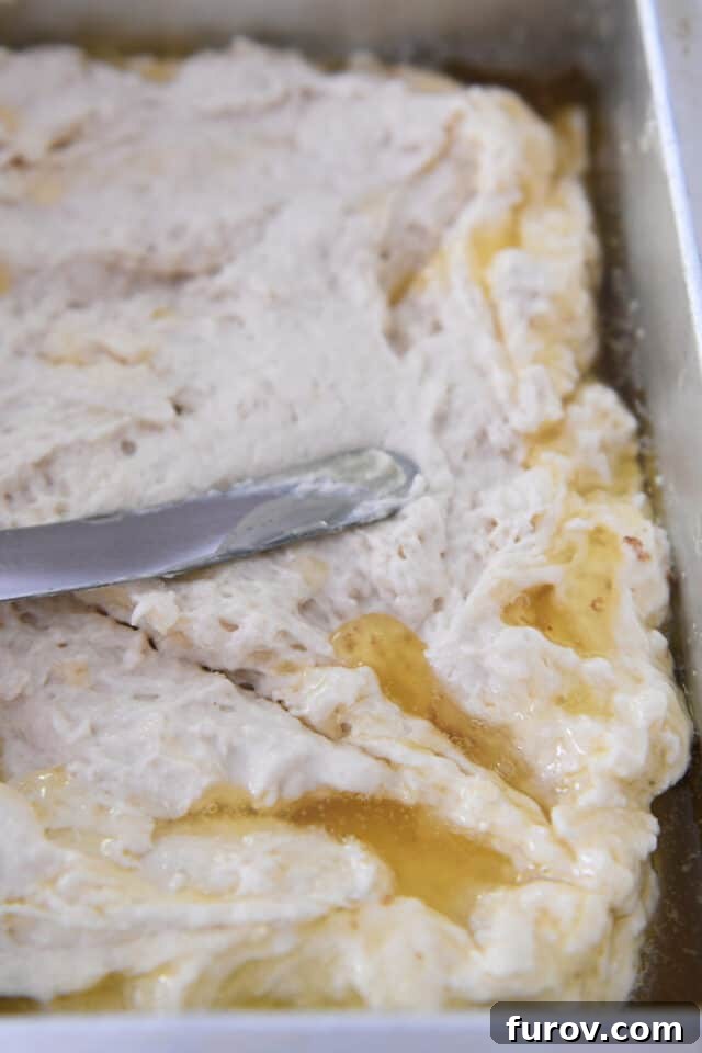A spatula gently spreading thick biscuit batter into a square baking pan, where a generous amount of melted butter already pools at the bottom and sides.