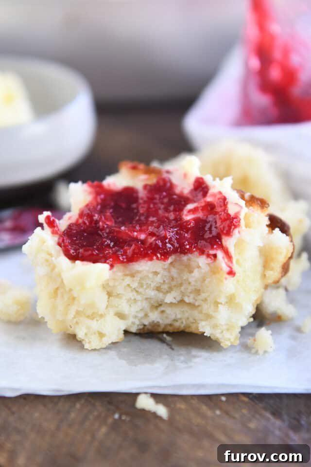 A close-up of a Butter Swim Biscuit with a bite taken out, generously smothered with melting butter and vibrant raspberry jam, highlighting its delicious texture.