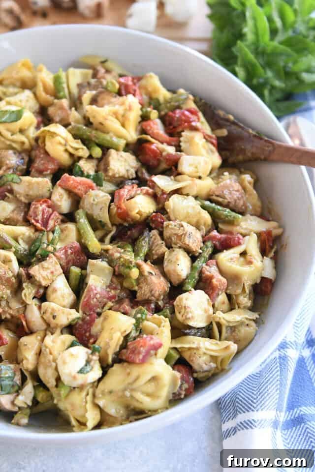 Tangy Pesto Balsamic Chicken Tortellini Medley 2 A beautifully presented white bowl of tortellini salad featuring fresh mozzarella, grilled chicken, roasted red peppers, and cherry tomatoes, with a wooden spoon resting beside it.