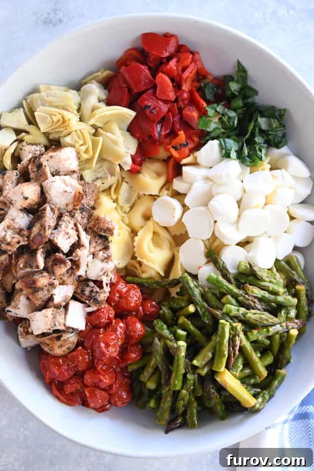 Tangy Pesto Balsamic Chicken Tortellini Medley 4 A vibrant collection of fresh ingredients in a white bowl, including artichokes, roasted red peppers, fresh basil, mozzarella, asparagus, roasted tomatoes, grilled chicken, and tortellini, ready to be tossed.