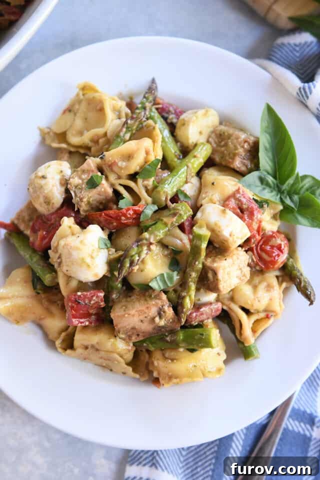 Tangy Pesto Balsamic Chicken Tortellini Medley 8 A white plate showcasing a generous serving of tortellini salad, adorned with fresh mozzarella balls, grilled chicken pieces, and a garnish of fresh basil.