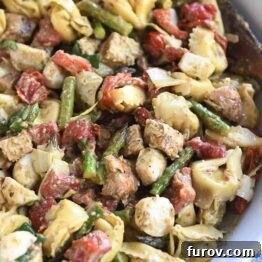 A white bowl with a wooden spoon featuring a vibrant tortellini salad with mozzarella, chicken, roasted red peppers, and tomatoes.