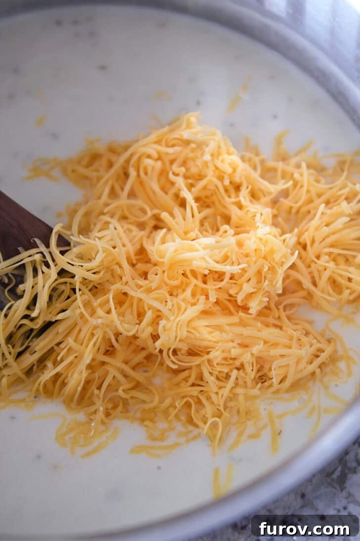 A creamy white sauce in a pot, topped with a generous pile of shredded sharp cheddar cheese, ready to be melted.