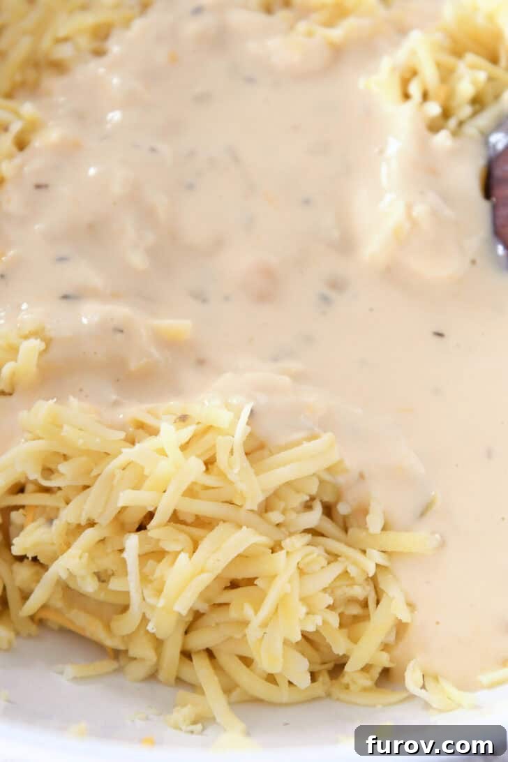 Top-down view of rich shredded cheese being stirred into a creamy white sauce in a white mixing dish, creating a luscious blend.