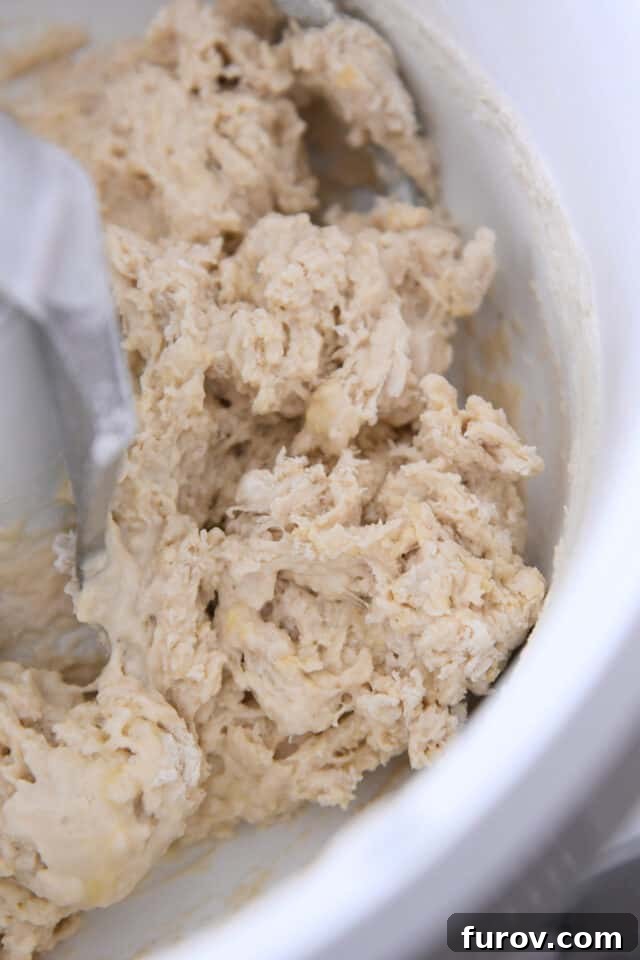 Initial shaggy and messy dinner roll dough in a white mixing bowl with a dough hook, indicating the early stage of mixing.