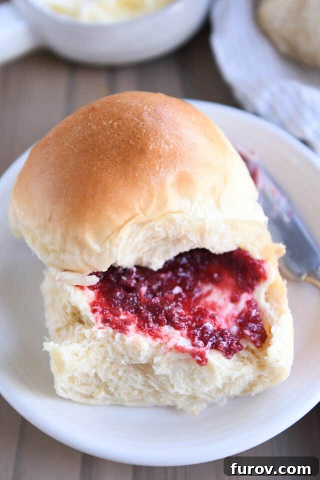 A soft dinner roll sliced open on a white plate, generously spread with butter and a dollop of fruit jam, inviting a delicious bite.