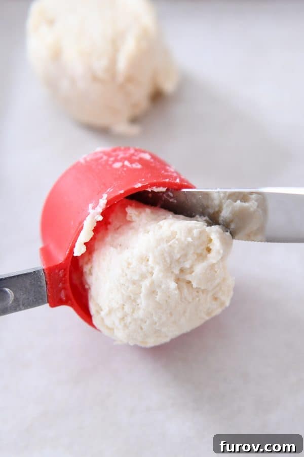 A butter knife assisting in scooping sticky dough from a measuring cup onto a baking sheet, highlighting the practical technique for drop biscuits.