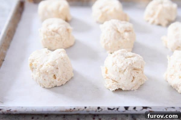 Mounds of unbaked buttermilk drop biscuit dough neatly arranged on a parchment-lined baking sheet, ready for the oven.