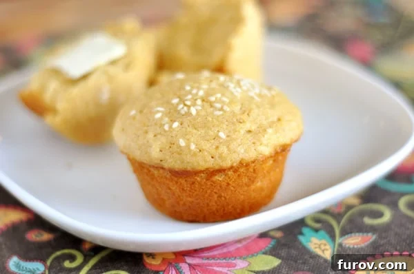 A beautifully golden whole wheat dinner muffin topped with sesame seeds, presented alongside another muffin cut in half on a white plate, highlighting the delicious texture within.