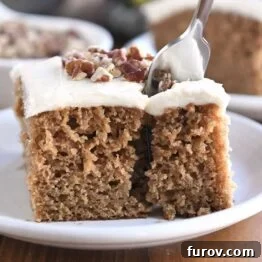 Fork taking bite of frosted applesauce spice cake on white plate.