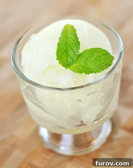 A small glass goblet filled with vibrant yellow lemon ice, garnished with fresh green mint leaves, sits on a white surface, evoking a sense of cool refreshment.