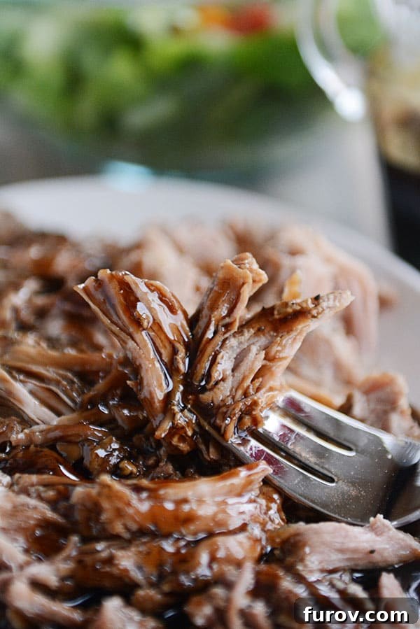 A close-up of a fork holding a succulent piece of glazed shredded pork, with a full plate and a fresh salad blurred in the background, highlighting the dish's appeal.