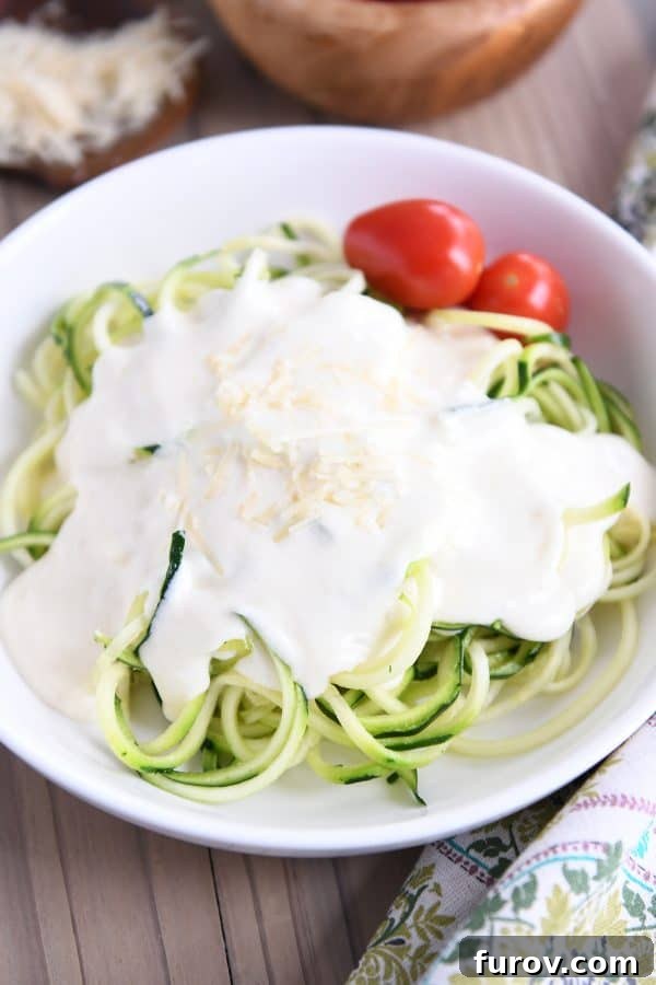 Creamy homemade Alfredo sauce generously coats vibrant green zucchini noodles (zoodles) in a white bowl, offering a lighter alternative to traditional pasta.