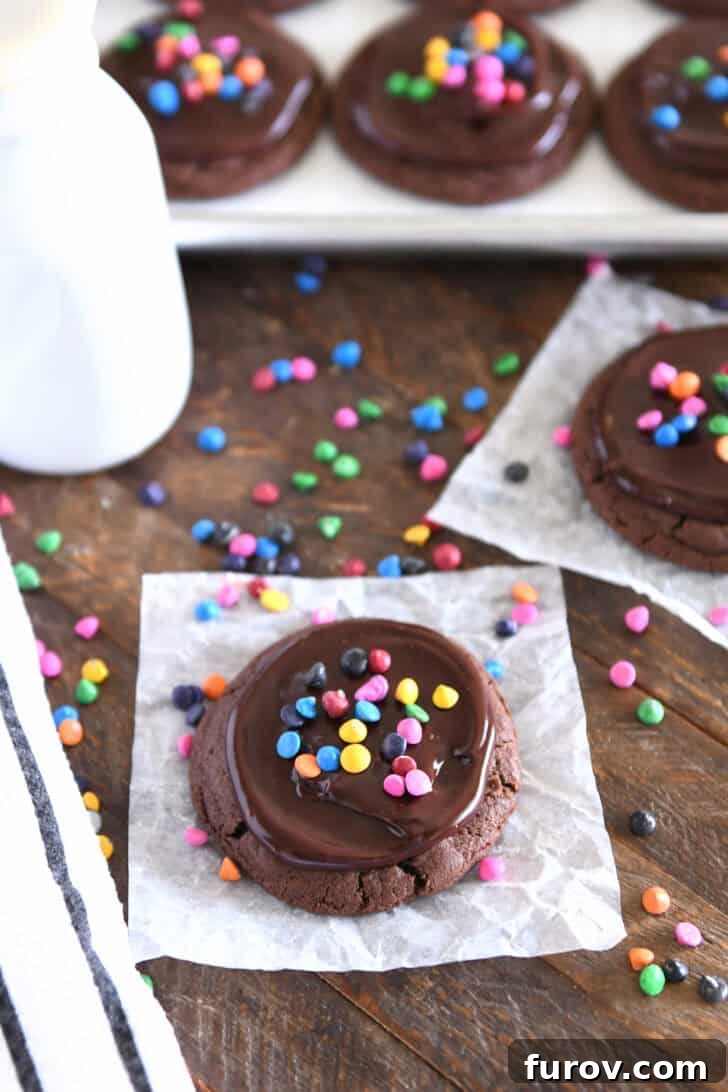 Chocolate cookie topped with ganache and rainbow chips on white parchment paper with rainbow chips sprinkled around and several cookies in the background.