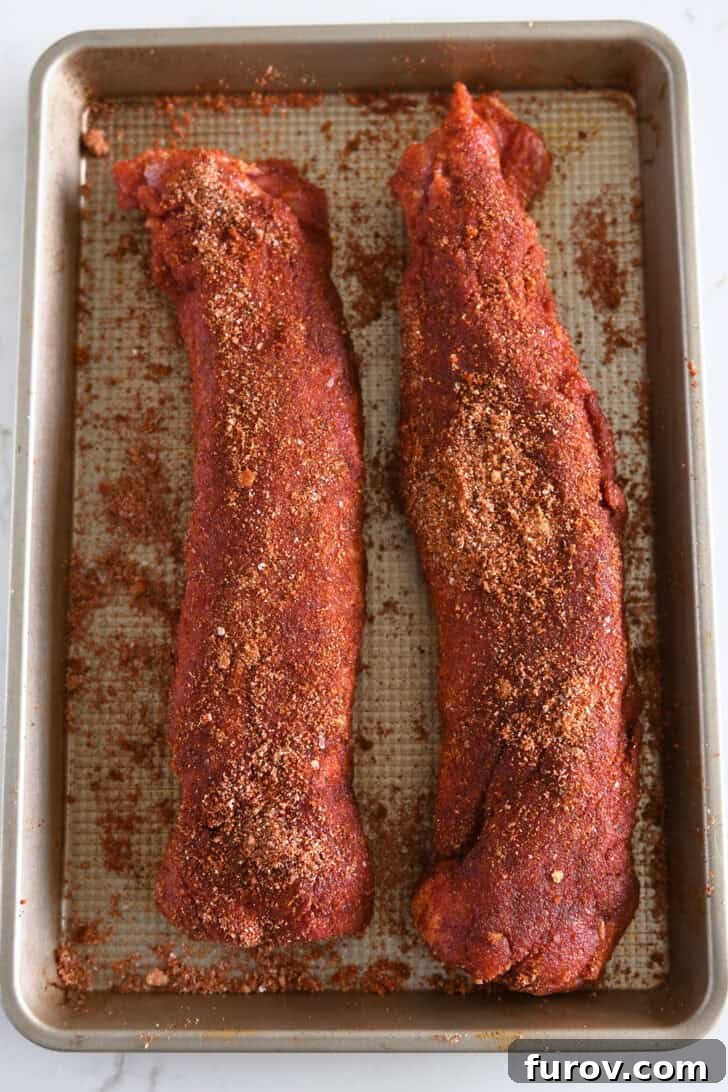 Speedy and Succulent Air Fryer Pork Tenderloin 4 Two raw pork tenderloins generously coated with the spice rub on a gold baking sheet, ready for air frying.