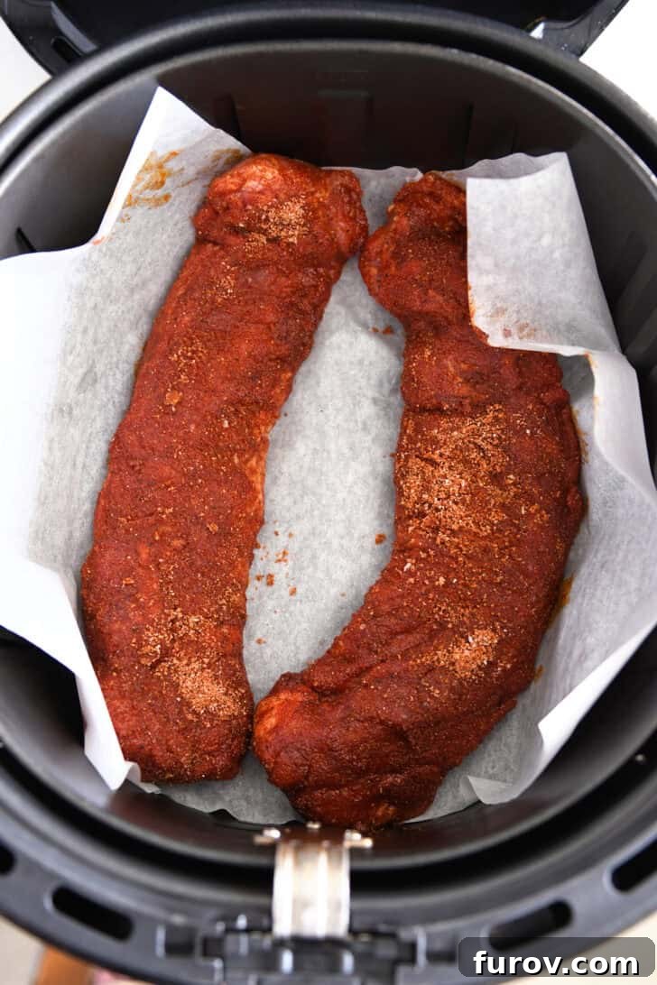 Speedy and Succulent Air Fryer Pork Tenderloin 5 Two raw, spice-crusted pork tenderloins placed on parchment paper inside an air fryer basket, ready to cook.