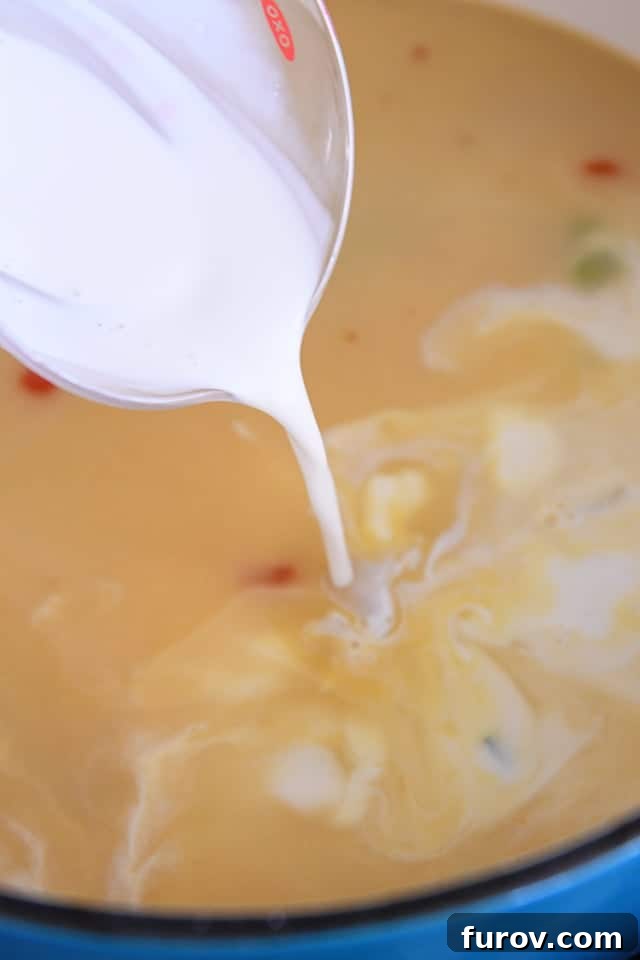 A steady stream of half-and-half being poured into a pot, mixing with chicken broth and aromatic vegetables, to create a creamy chowder.
