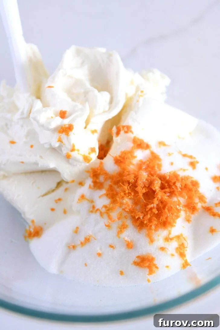 Glass bowl filled with cream cheese, sugar, and fresh orange zest, prepared for mixing.