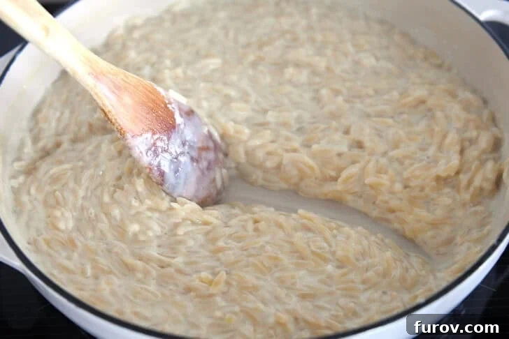 Wooden spoon scraping path through creamy orzo in white pan.