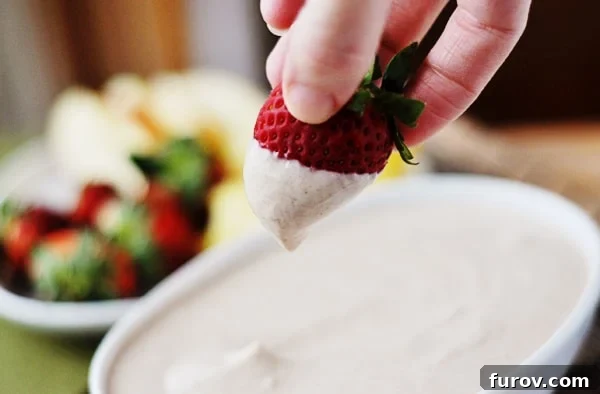 A perfectly half-dipped strawberry poised above a pristine white bowl brimming with creamy honey-yogurt fruit dip, highlighting its smooth texture and inviting appeal.