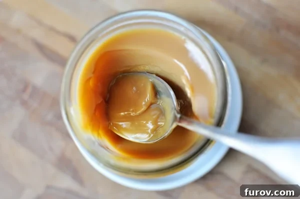 Whip Up Your Own Dulce de Leche 2 A top-down view of a spoon scooping rich, golden dulce de leche from a clear glass mason jar, showcasing its creamy texture.