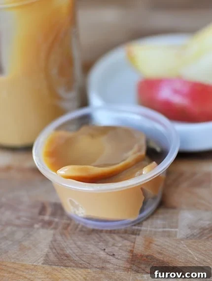 Whip Up Your Own Dulce de Leche 5 A small plastic container of creamy dulce de leche sits in the foreground, with a larger glass mason jar of dulce de leche and fresh apple slices blurred in the background, ready for dipping.