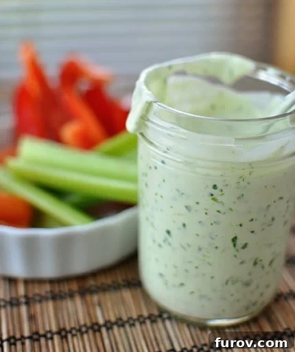 Glass mason jar full of homemade ranch dressing, with a bowl of fresh cut vegetables behind it. A perfect scene for a healthy snack option.