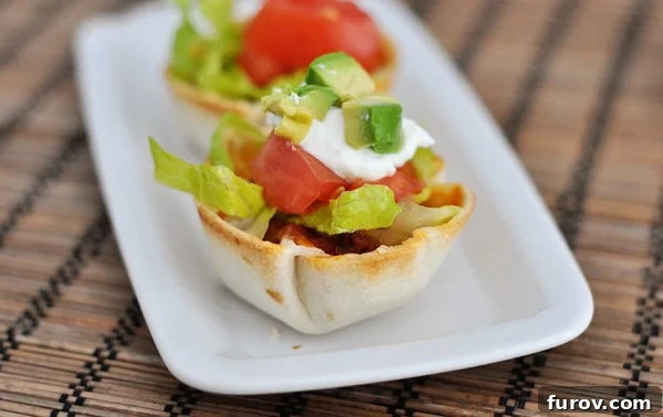 Two vibrant mini taco cups, generously topped with crisp lettuce, juicy diced tomatoes, creamy sour cream, and fresh avocado, arranged on a white serving platter.