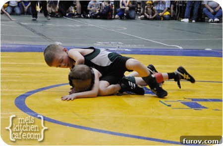 A young wrestler in a blue singlet, focused during a match on the mat, with spectators in the background, illustrating sideline etiquette.