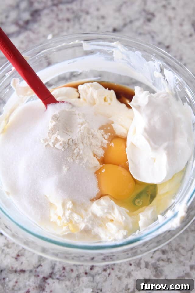 Fresh ingredients for cheesecake batter including eggs, granulated sugar, cream cheese, sour cream, and flour, artfully arranged in a clear glass bowl, ready for mixing.